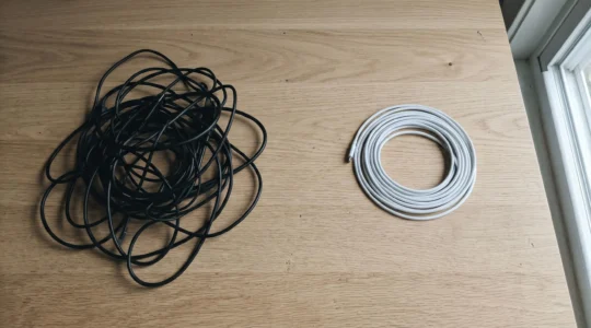 Sur une table en bois clair, fils électriques noirs emmêlés à gauche, câble blanc enroulé à droite, lumière douce venant de la droite